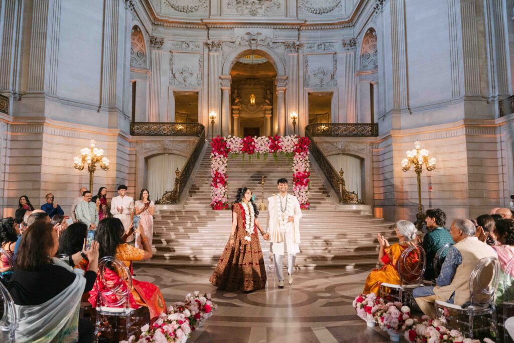Indian wedding couple walking down the grand staircase at San Francisco City Hall during ceremony recessional with guests seated on both sides